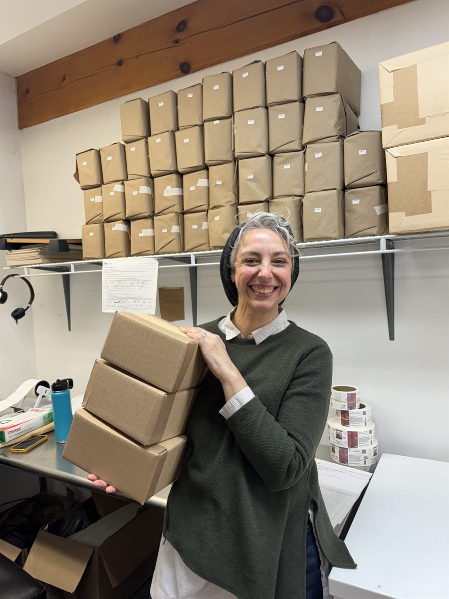 Eliza carrying finished shipping boxes, shelves of wrapped orders behind her